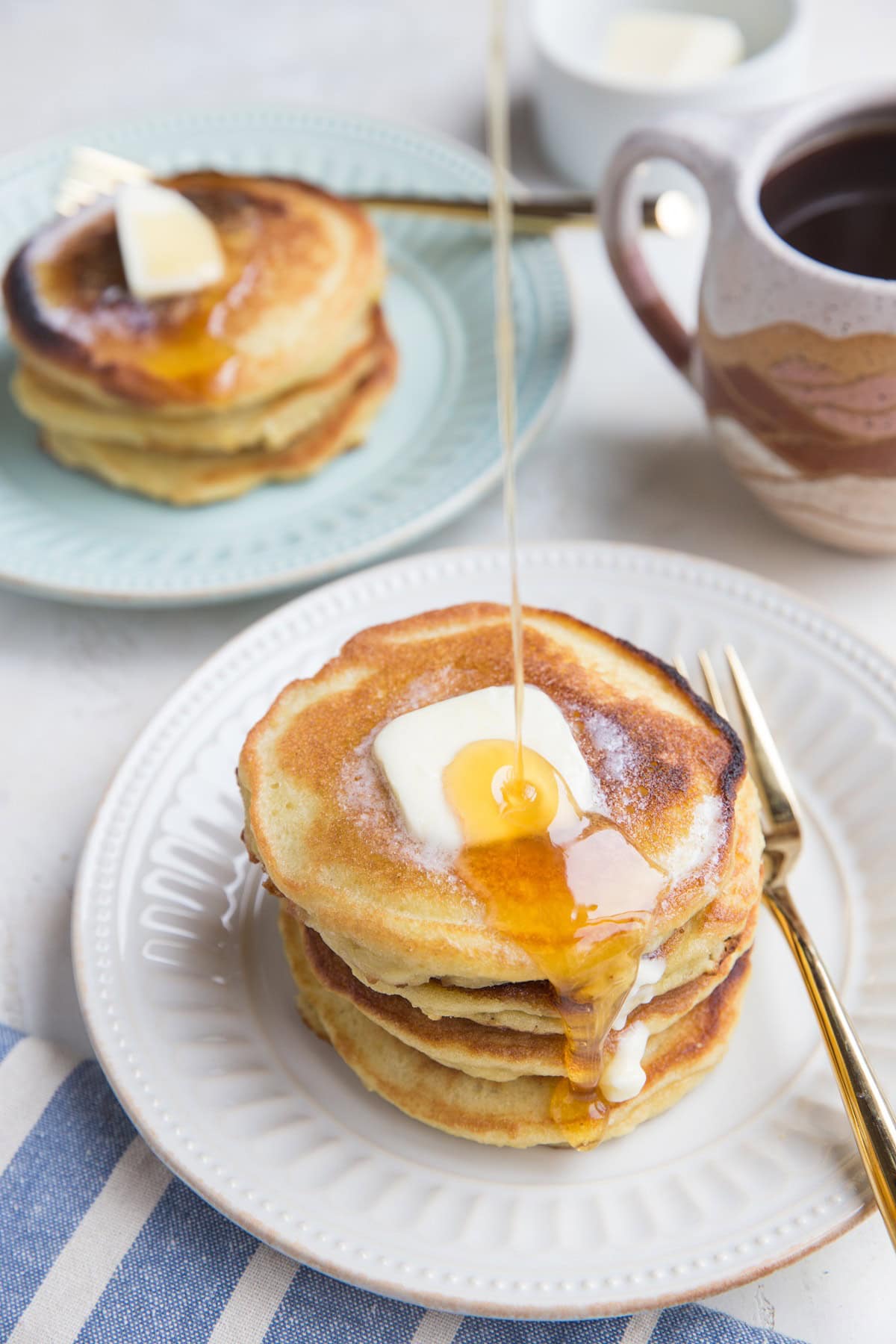 Two plates of pancakes with honey being poured on top of one of the stacks.