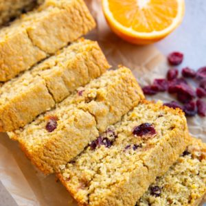 Loaf of cranberry orange bread cut into slices with a fresh orange and dried cranberries in the background.