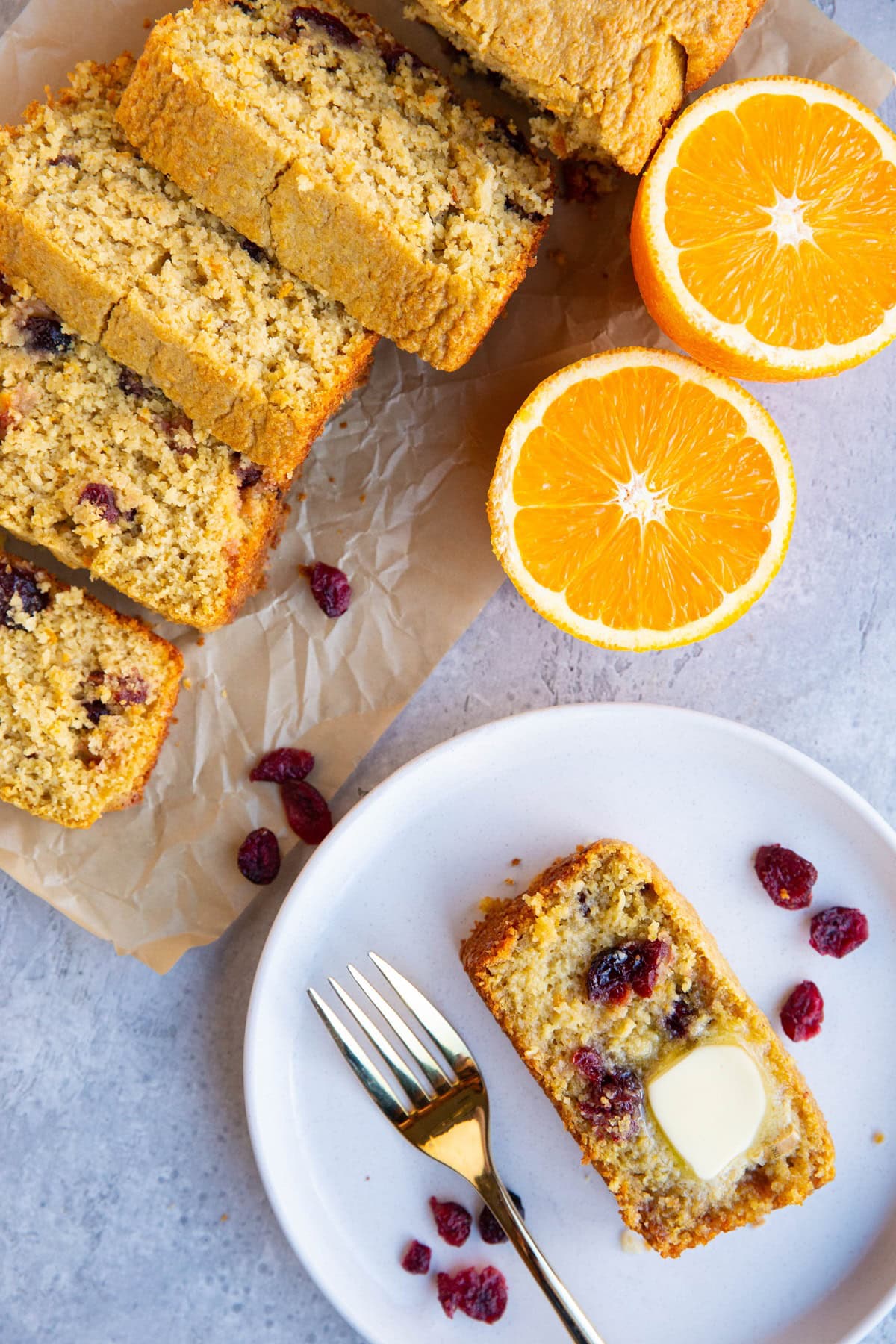 Slice of cranberry orange bread on a plate with the rest of the loaf next to it cut into slices and a fresh orange to the side.