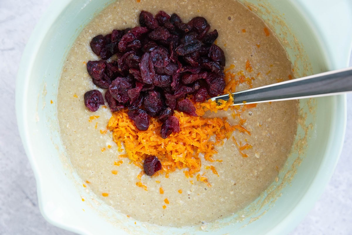 Muffin batter in a mixing bowl with orange zest and dried cranberries on top, ready to be mixed in.