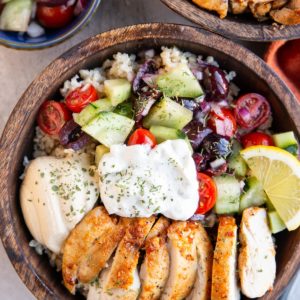 Two wooden bowls full of chicken , hummus, tzatziki sauce, and cucumber tomato salad, ready to serve.