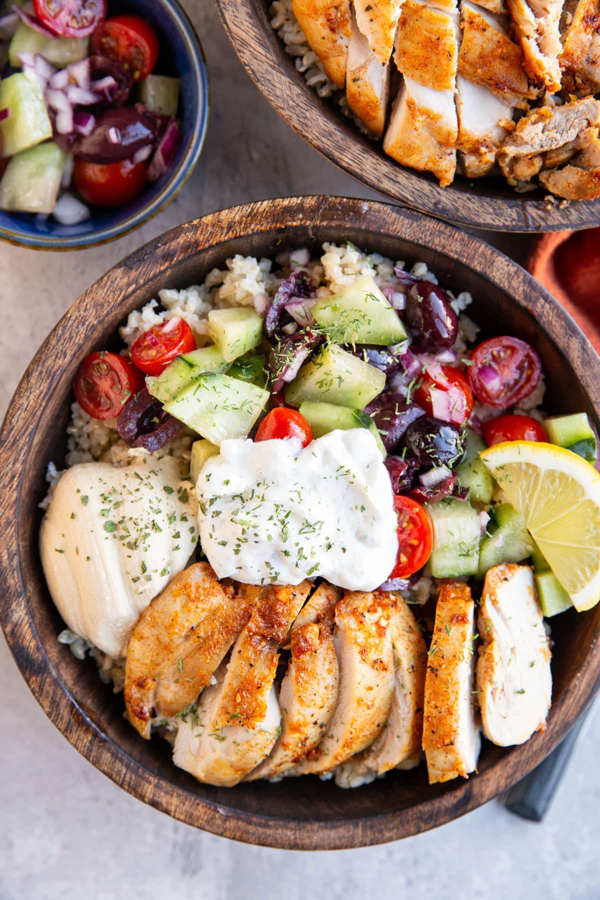 Two wooden bowls full of chicken , hummus, tzatziki sauce, and cucumber tomato salad, ready to serve.