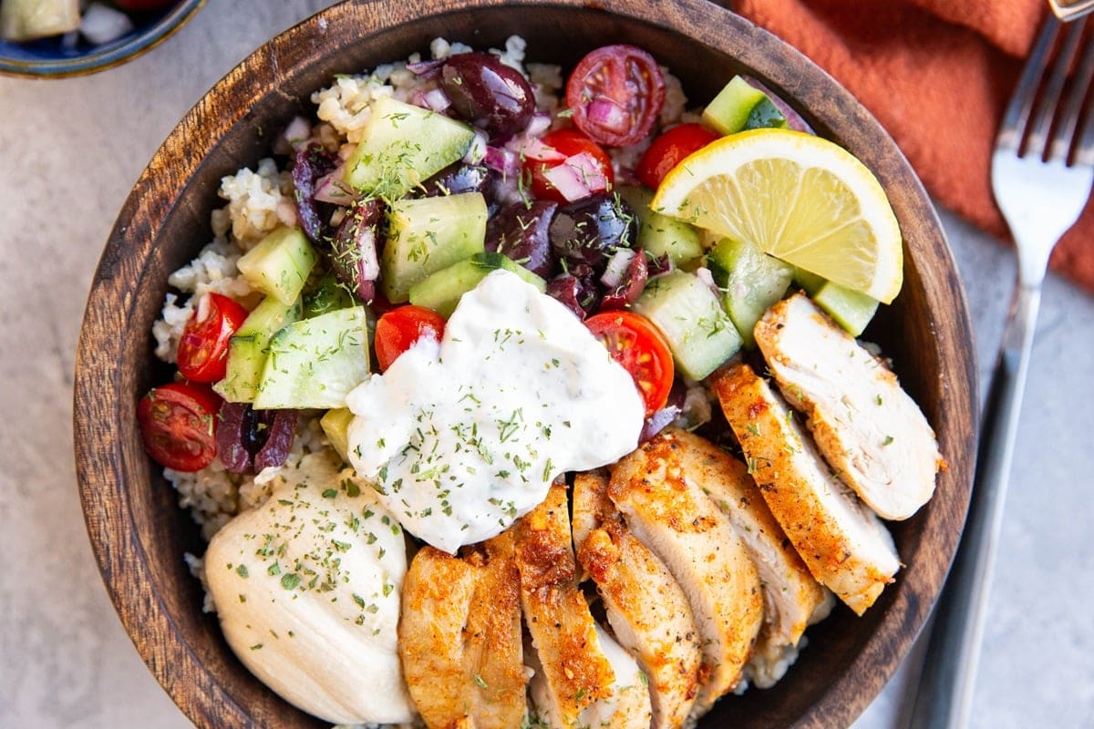 Big wooden bowl full of chicken, cucumber salad, tzatziki sauce, hummus, and brown rice. A fork and a napkin to the side.