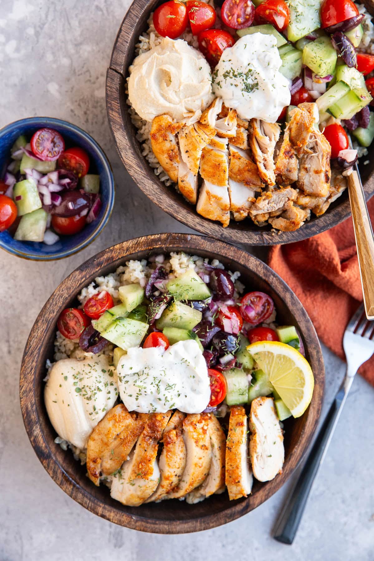 Two wooden bowls full of brown rice, baked chicken, tomato salad, and hummus. A bowl of tomato salad to the side