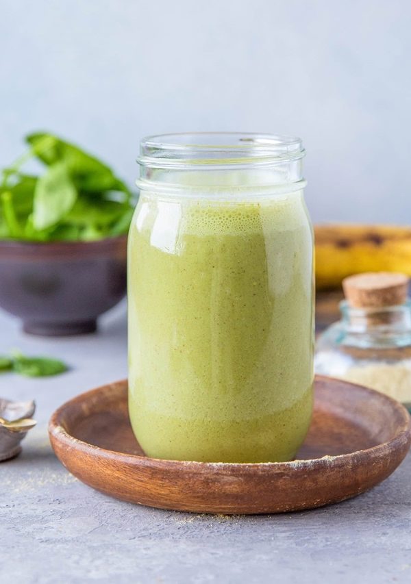 Green smoothie in a glass on top of a wooden plate with fresh spinach and banana in the background.