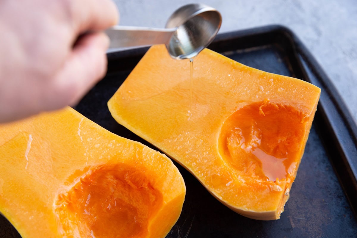 Hand pouring avocado oil over butternut squash halves.