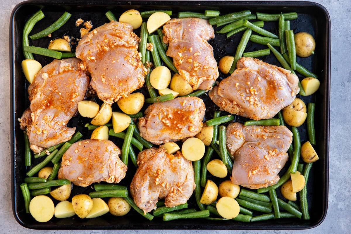 Baking sheet with green beans, potatoes, and raw chicken thighs on top, ready to go into the oven.