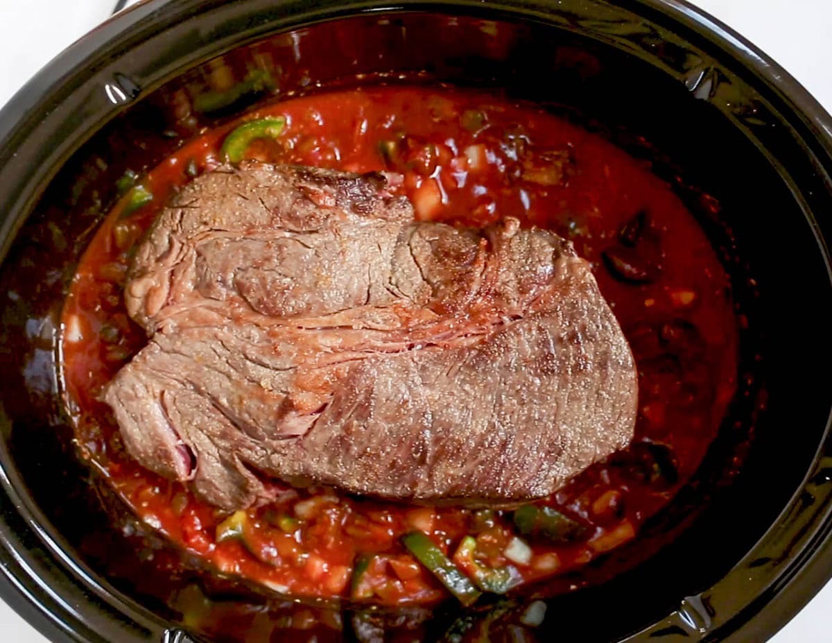 Big chunk of meat sitting in tomato pepper sauce in a slow cooker, ready to cook.