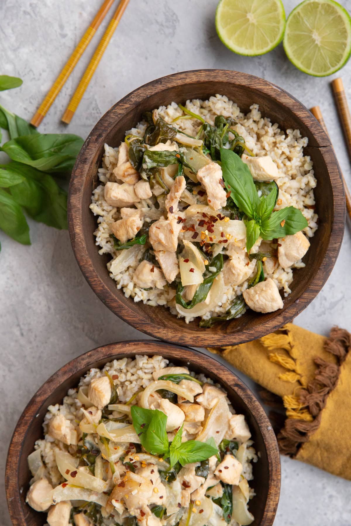 Two wooden bowls full of brown rice and coconut lime chicken with fresh basil and red pepper flakes on top. Chop sticks and lime wedges to the side.