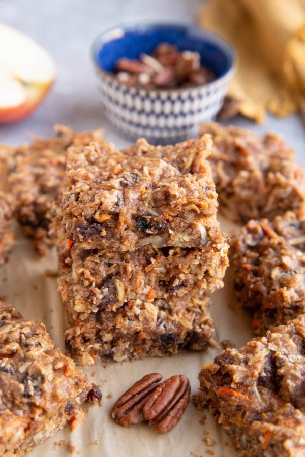 Stack of carrot cake breakfast bars on a sheet of parchment paper with the pecans in the background.