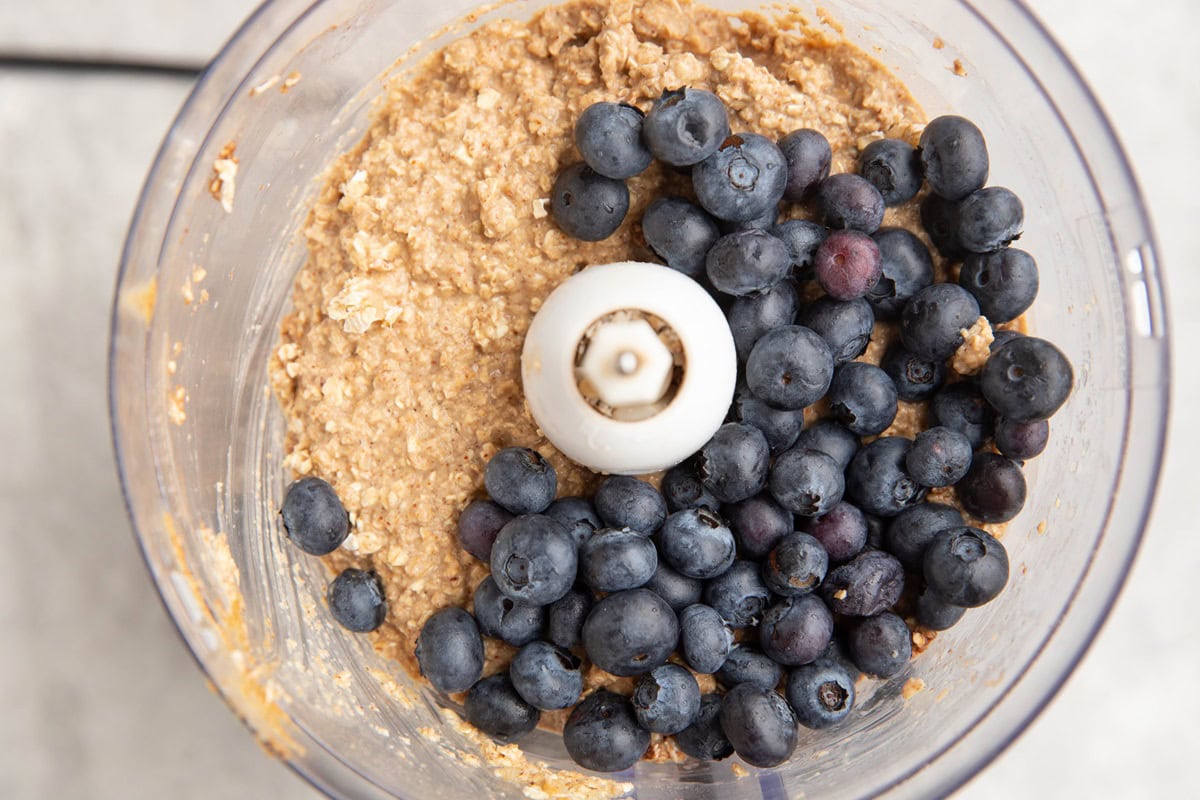 Food processor with oatmeal mixture and blueberries on top, ready to be mixed in.