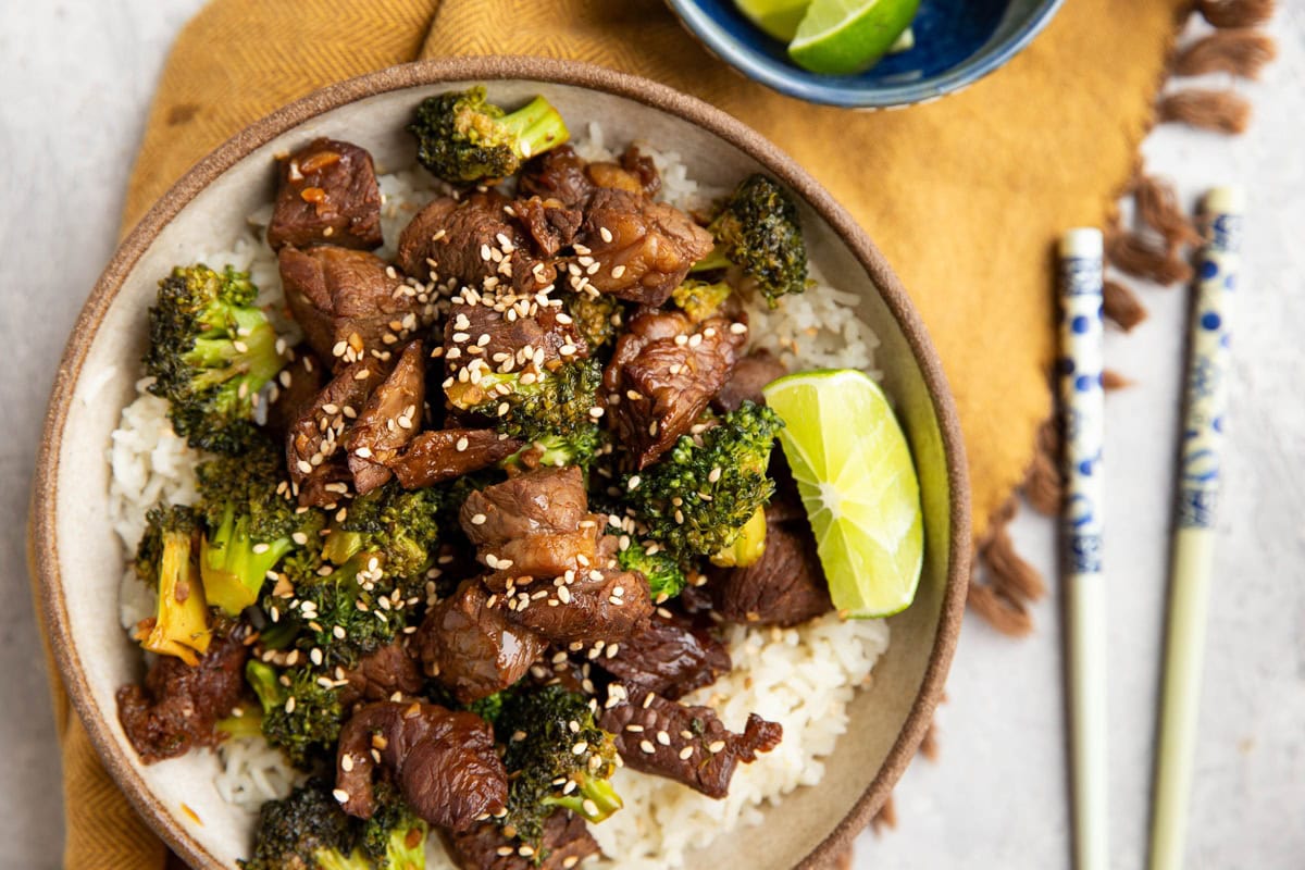 Broccoli and beef in a large bowl on top of rice with chopsticks and a golden napkin.