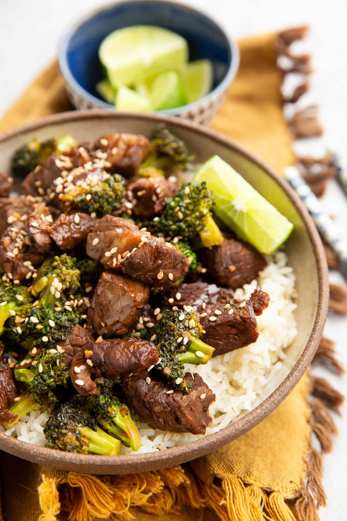 Broccoli and beef in a large bowl on top of white rice with wedges of fresh limes in the background.