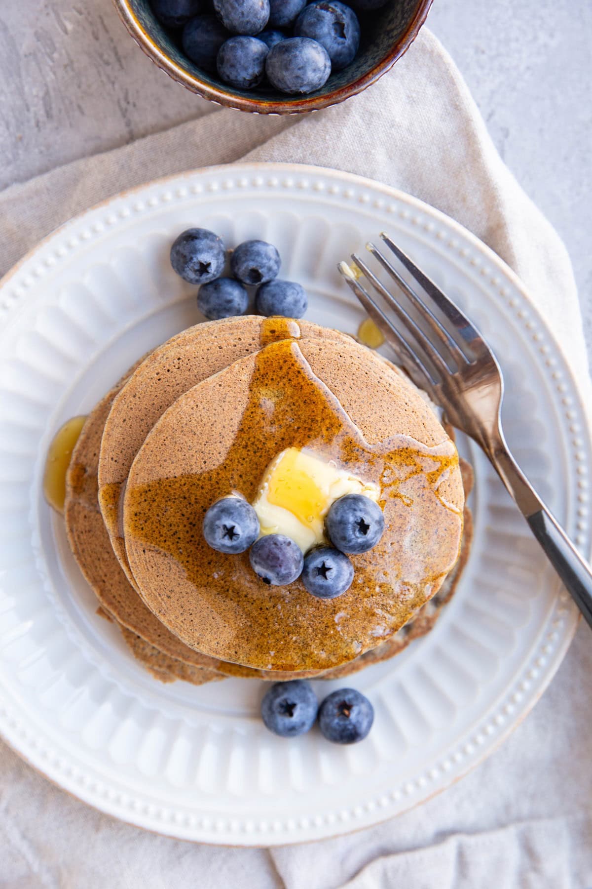 Stack of buckwheat pancakes on a plate with butter and blueberries on top, drizzled with pure maple syrup.