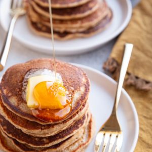 Stack of pancakes with melted butter on top with honey being poured over the top. A stack of pancakes in the background.