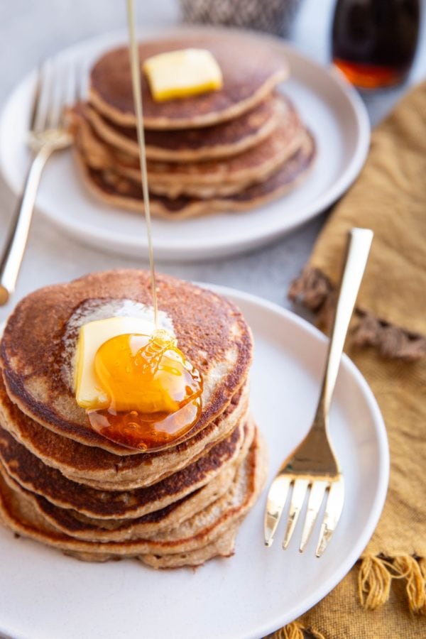 Stack of pancakes with melted butter on top with honey being poured over the top. A stack of pancakes in the background.