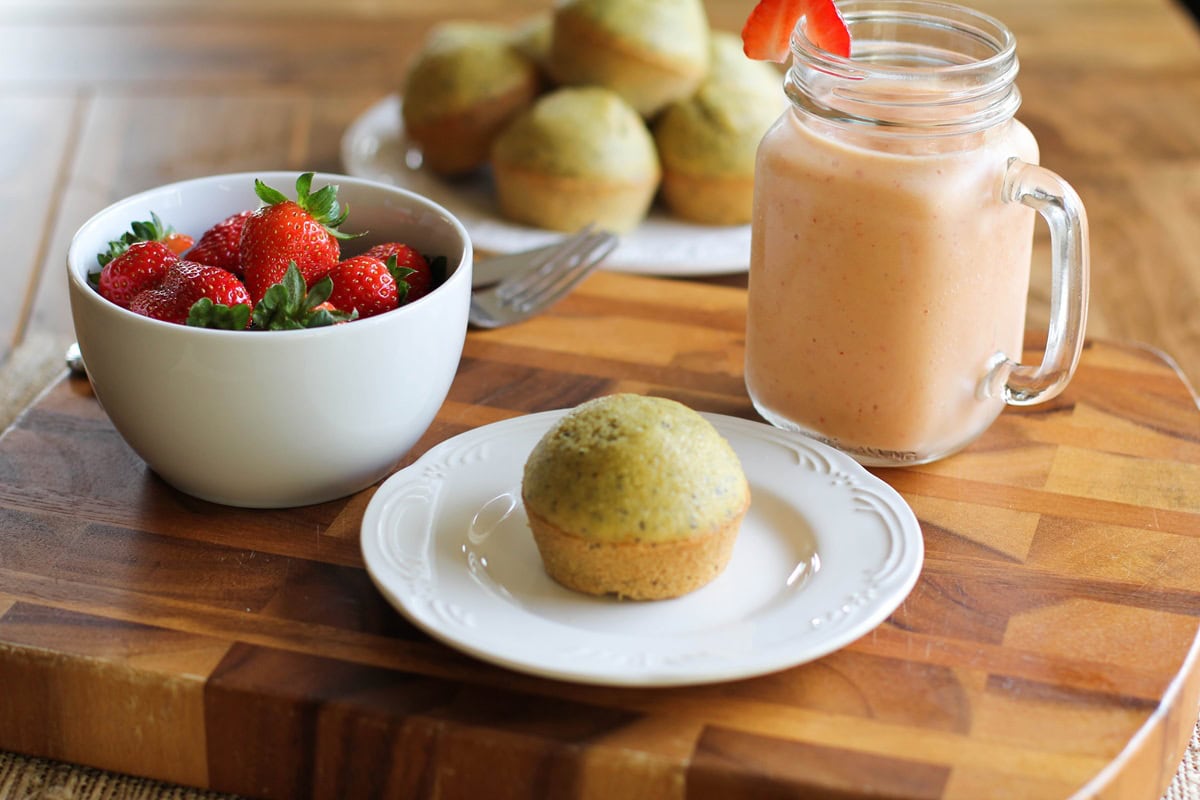 Wooden tray of gluten-free lemon poppy seed muffins with a bowl of fruit and a smoothie to the side.