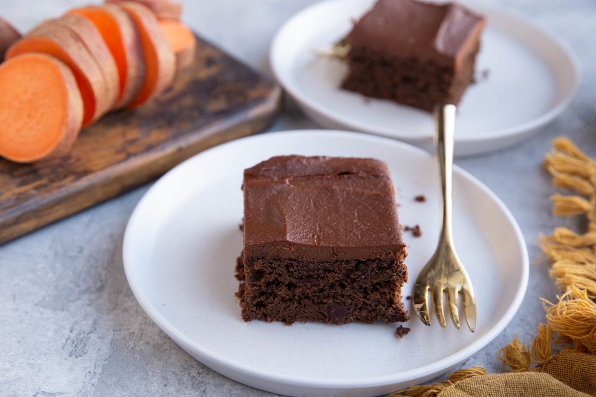 Two plates with slices of chocolate cake with chocolate frosting and a chopped up sweet potato in the background.