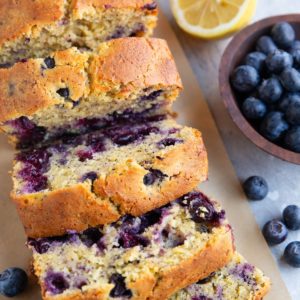 Loaf of lemon blueberry bread cut into slices with a bowl of blueberries and cut lemon in the background.