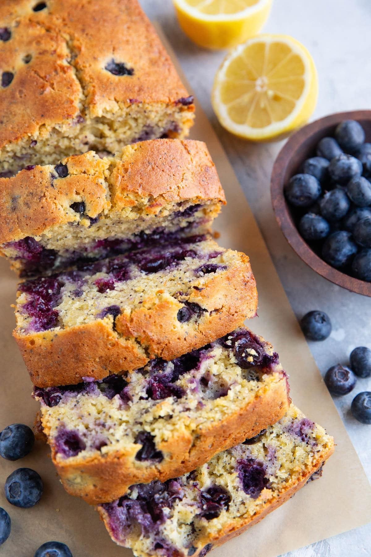 Loaf of lemon blueberry bread cut into slices with a bowl of blueberries and cut lemon in the background.