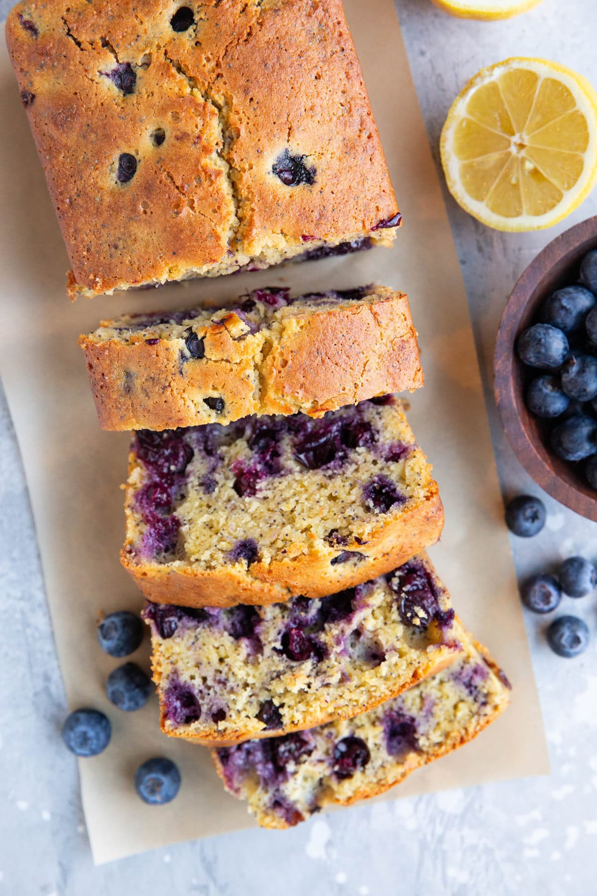 Loaf of blueberry lemon bread cut into slices with a blue of blueberries and fresh lemons to the side.