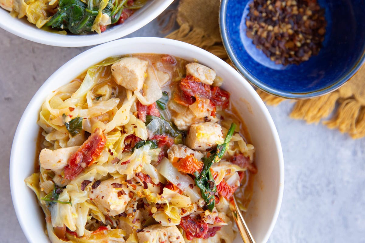 Sun-dried tomato chicken and cabbage in two bowls with red pepper flakes in a small bowl.