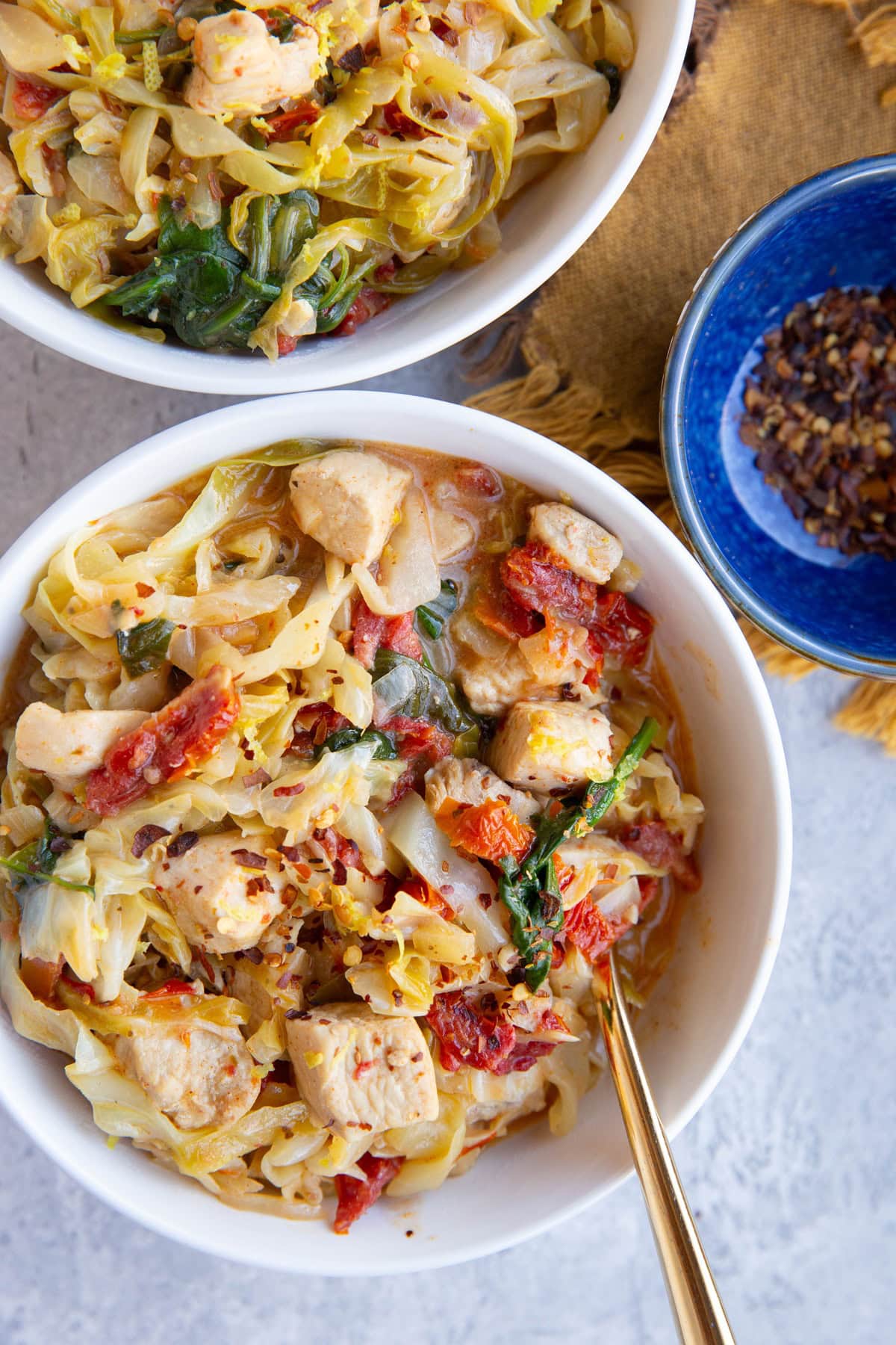 Two big white bowls full of chicken cabbage and creamy sauce. A bowl of red pepper flakes to the side.