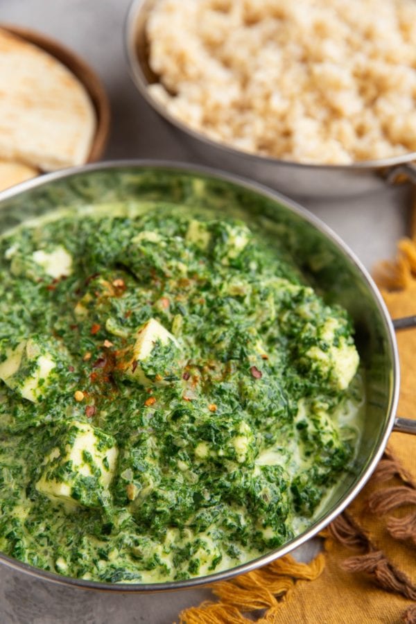 Indian dish full of saag paneer with a dish of brown rice in the background.